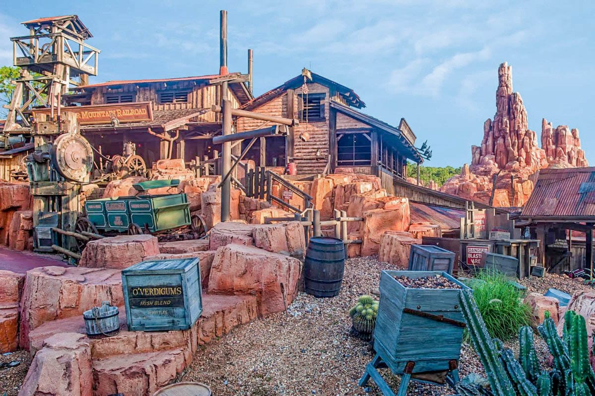 Old Western-themed Big Thunder Mountain Railroad station with red rocks, wooden buildings, barrels, and mining gear under a blue sky.