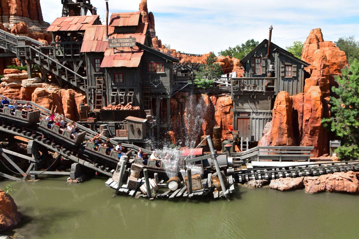 A Big Thunder Mountain Railroad train splashes through water by a Western mining building and red rock formations.