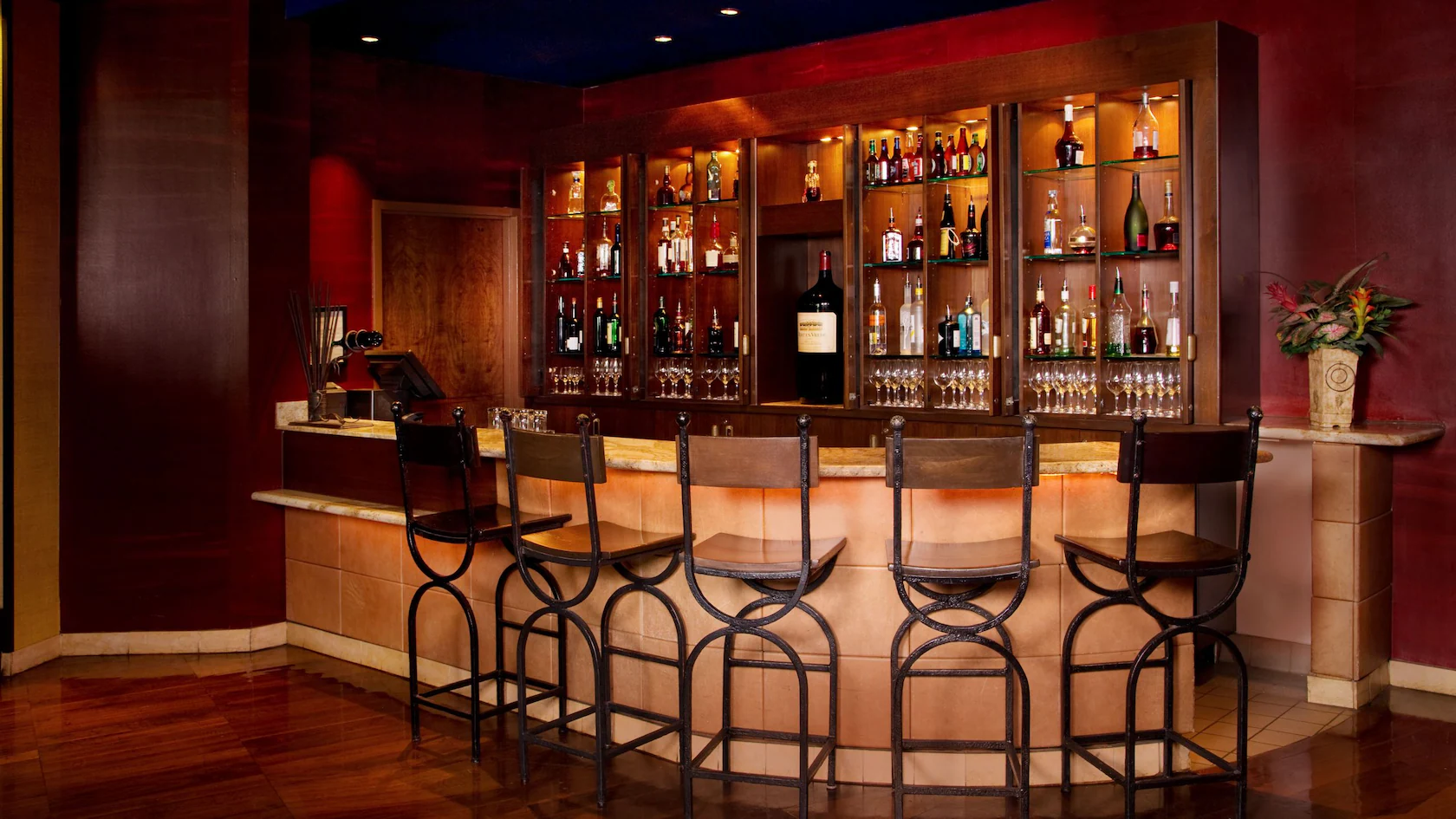 A modern wine bar with five tall chairs, a counter, and shelves of bottles and glasses against a red wall.