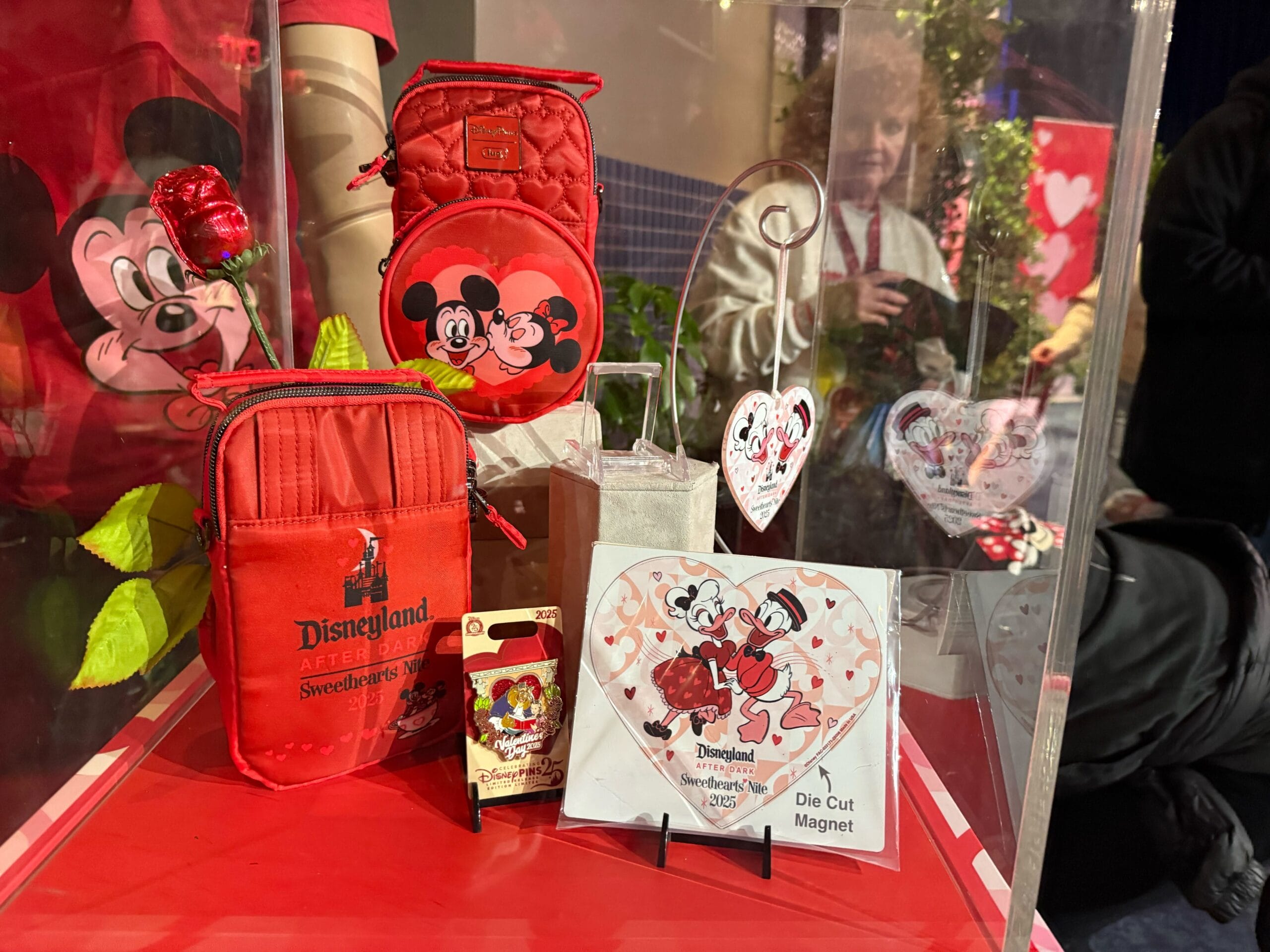 Display case featuring Sweethearts' Nite merchandise from Disneyland After Dark, with red-themed items like bags, a heart-shaped ornament, and a die-cut magnet starring Mickey and Minnie Mouse. Prices available upon request.