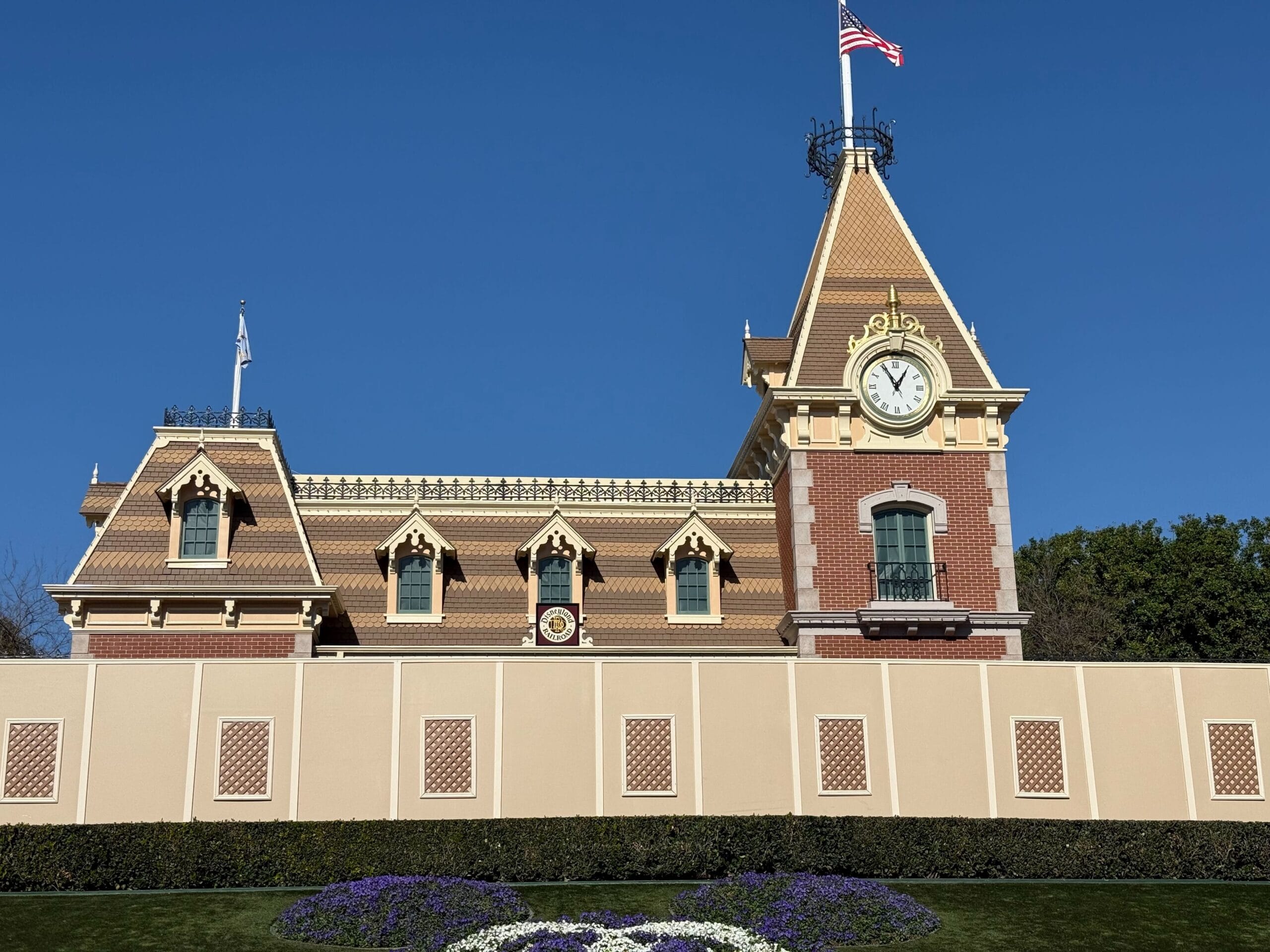 Clock tower of a red-brick train station building with decorative features, a flag on top, and a well-maintained garden with flower beds in front.