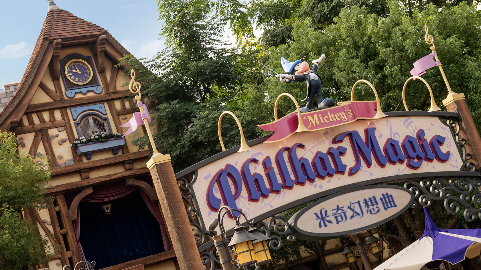 The entrance to Mickey's PhilharMagic attraction boasts a whimsical facade adorned with a clock tower and musical notes, while the sign, in both English and Chinese, welcomes visitors. Trees gracefully frame this enchanting Disney attraction.