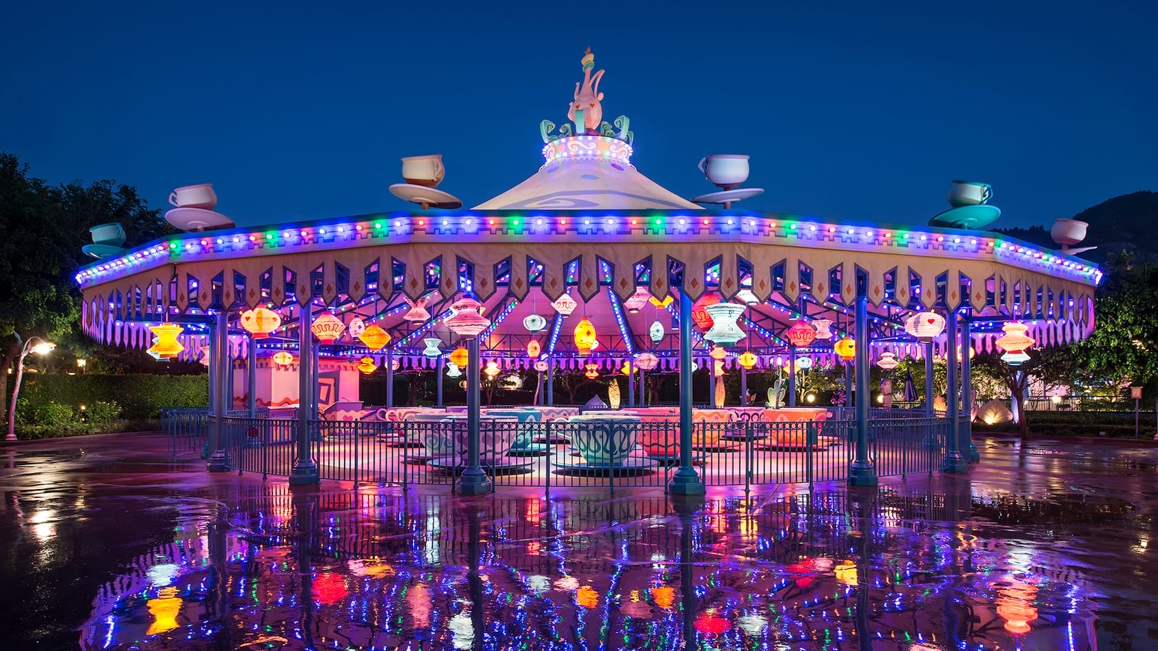 A brightly lit, circular amusement park ride with Mad Hatter Tea-themed teacup-shaped seats and a decorated canopy glowing at night. Reflections dance on the wet ground below, adding a touch of magic to the scene.