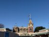 The Disneyland entrance showcases a Victorian-style train station flag waving proudly atop the clock tower, all set against a clear blue sky.