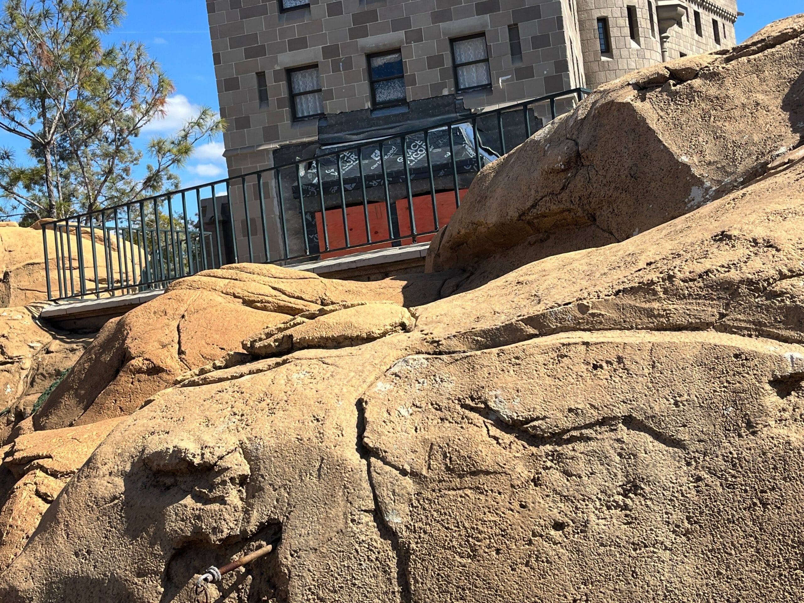 Amidst the large rocky terrain with a metal railing in the background, the Canada Pavilion at EPCOT features a building with a stone facade, perfectly set under a clear blue sky.