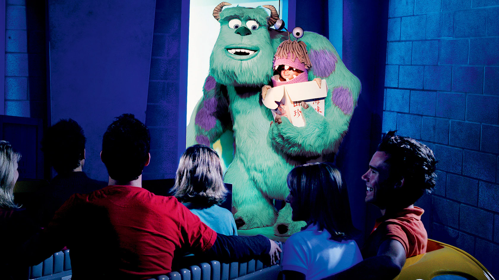A group of people on the ride watch animated characters, Mike & Sulley from Monsters, Inc., along with a furry monster and a child in costume, appear from a door illuminated by blue lighting.