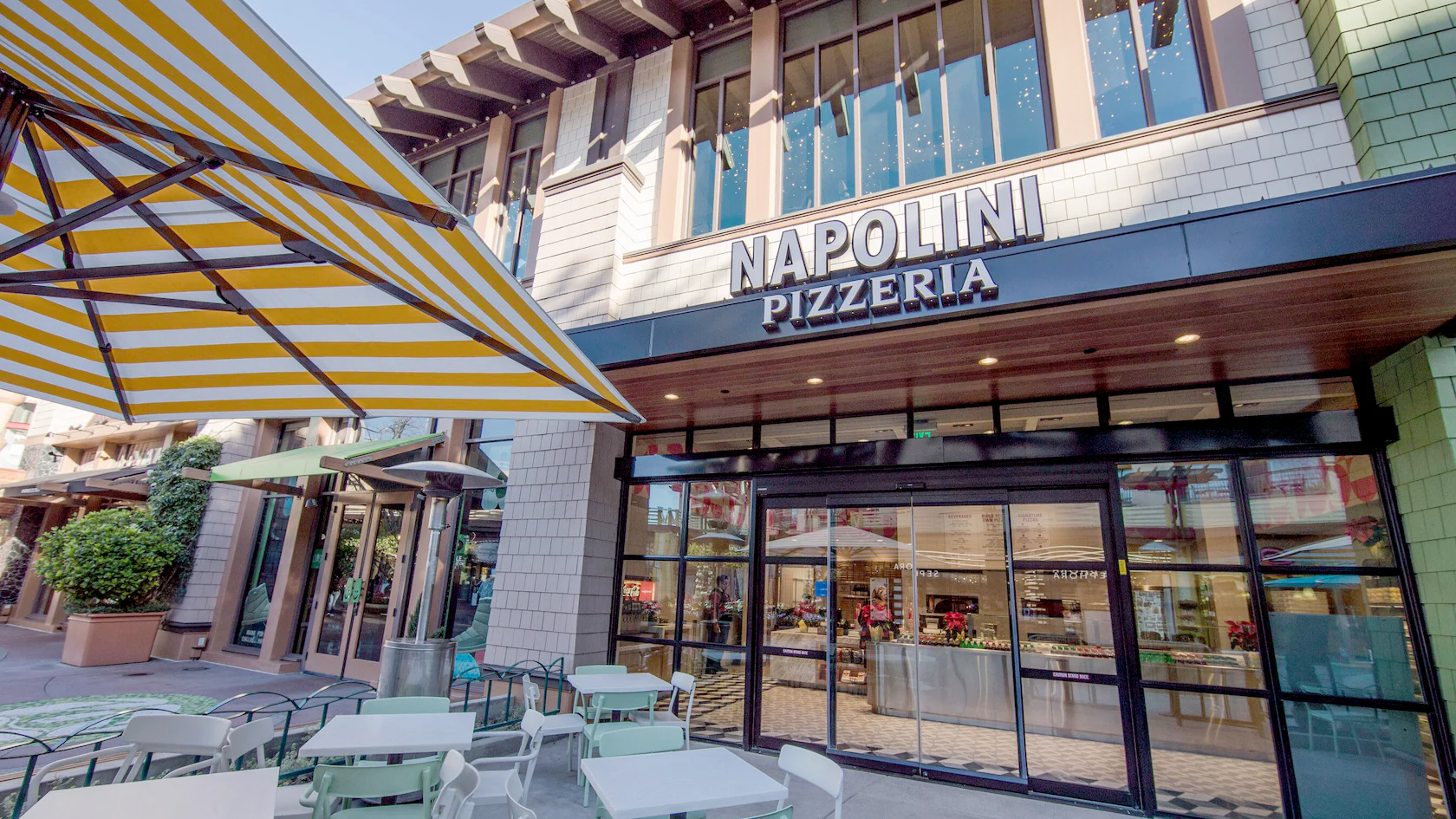 Outdoor seating at Napolini Pizzeria, an Italian restaurant, with tables under a yellow striped umbrella.