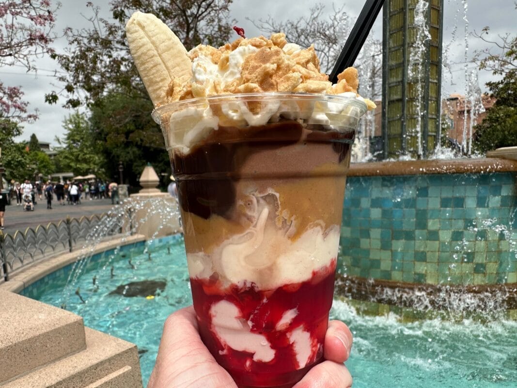 A towering PB&J Split dessert with layers of whipped cream, chocolate, peanut butter, banana, and red syrup is held in front of a fountain at Clarabelle's Ice Cream in Disney California Adventure.