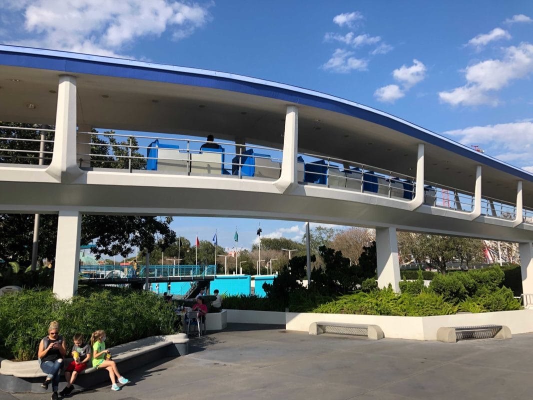 People sit on a bench beneath Tomorrowland's curved, elevated PeopleMover with blue railings on a sunny day.
