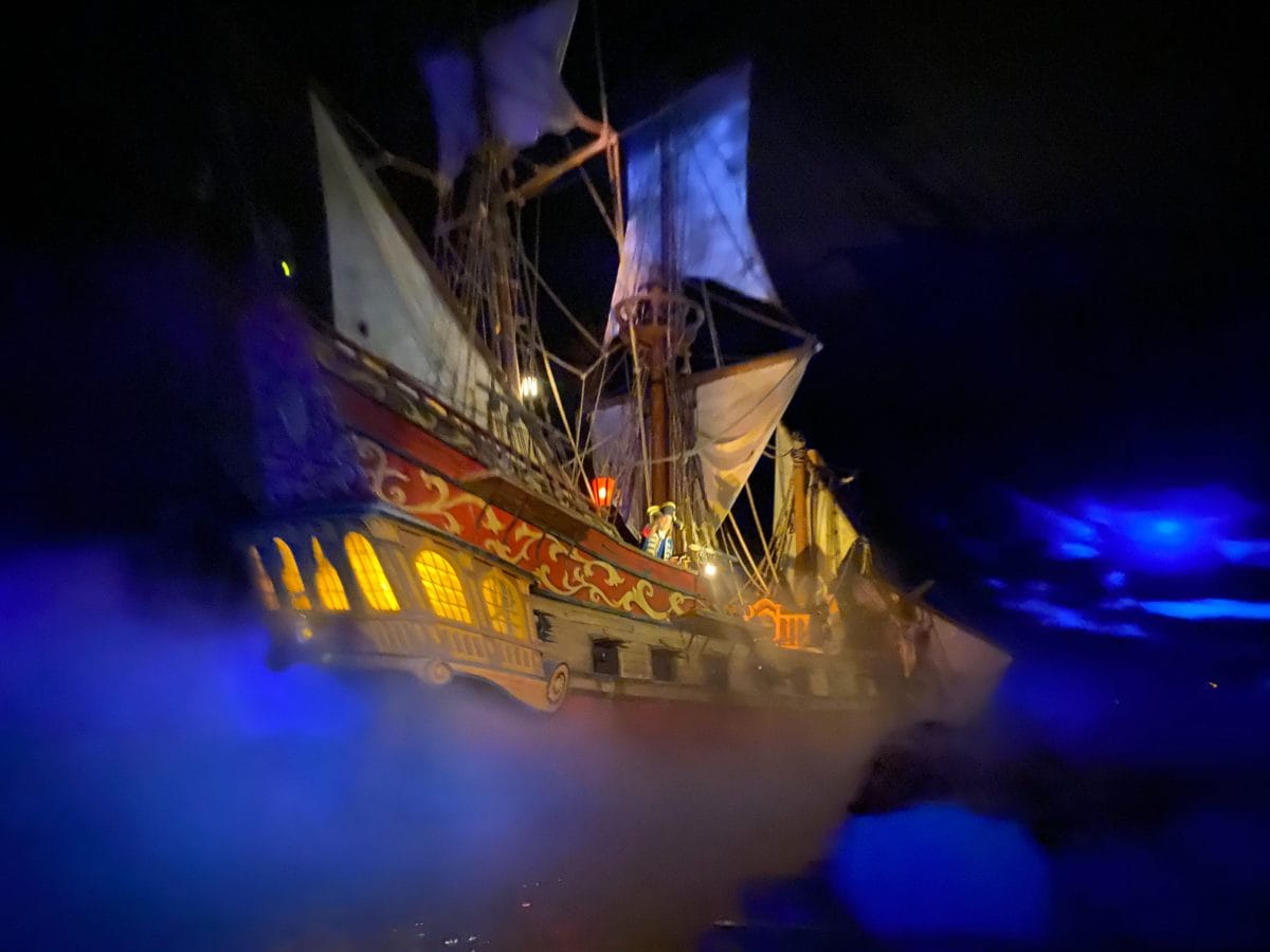 A Pirates of the Caribbean ship with glowing windows sails through mist in a dark, atmospheric blue-lit setting.