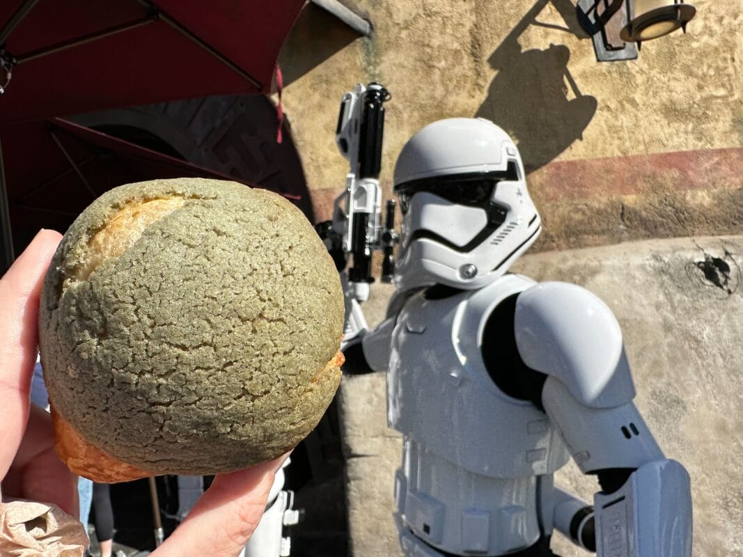 A person holds a greenish Polystarch Puffbread with a Stormtrooper figure in the background against a stone wall, reminiscent of Galaxy's Edge at Disneyland Park.