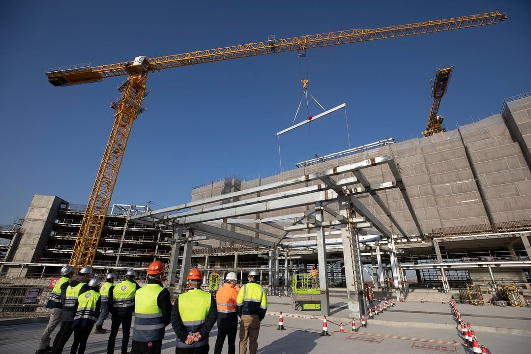 Construction site with workers and cranes lifting steel beams, surrounded by scaffolding as they lay the foundation for the Shanghai Disney Resort hotel structure. Workers in helmets and safety vests diligently observe the process under a clear blue sky.
