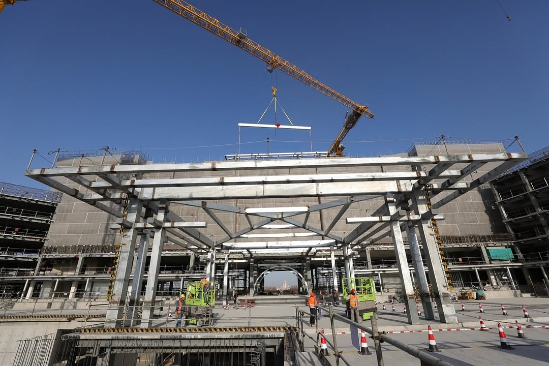 At a bustling construction site near Shanghai Disney Resort, a hotel structure is taking shape. Workers in safety gear collaborate as a crane expertly lifts a beam, assembling the steel frame under a clear blue sky, setting the stage for upcoming interior work.