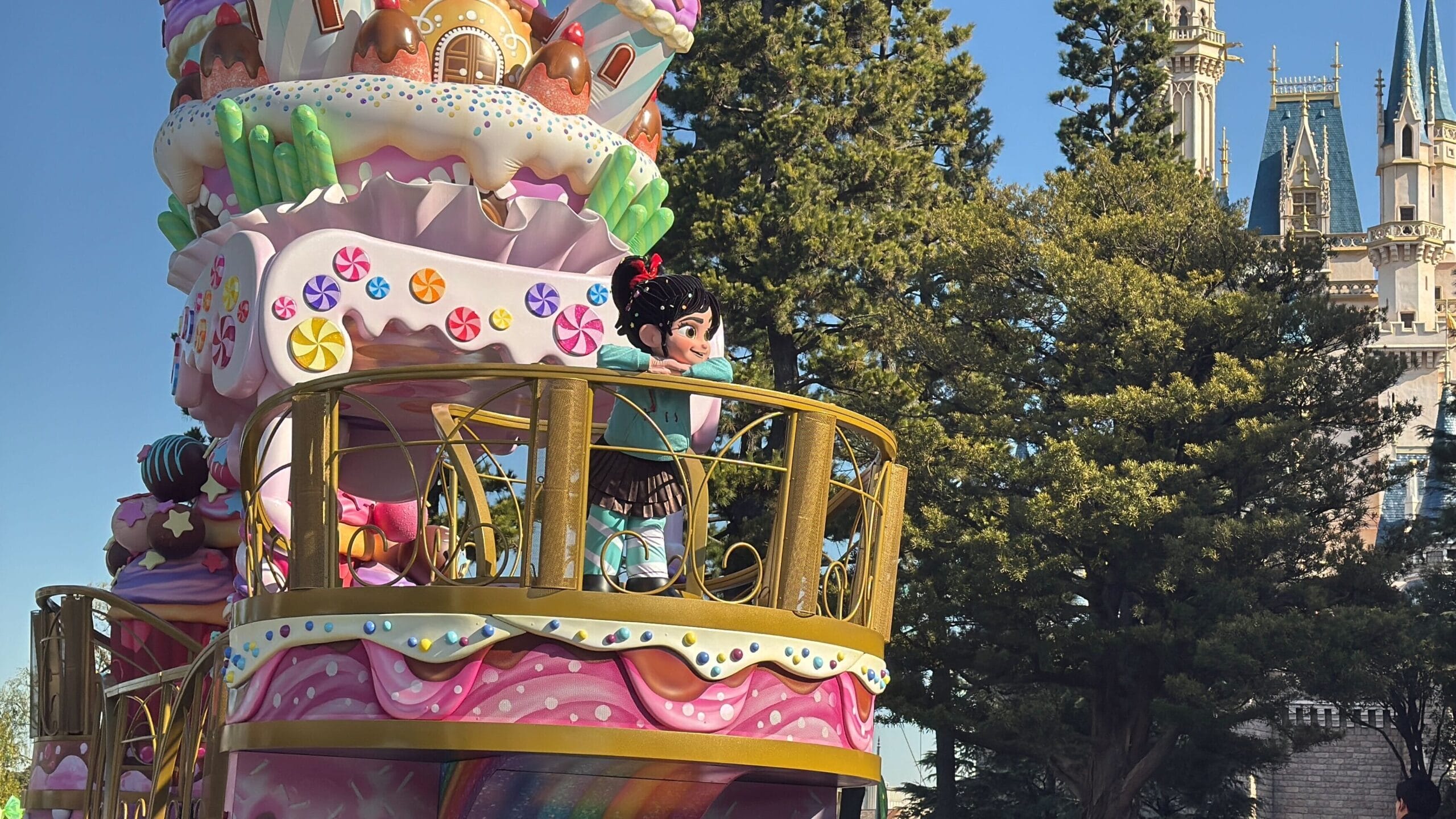 A colorful parade float bursts with a candy theme, featuring Vanellope in her iconic green and black outfit, set against the backdrop of a large castle and whimsical trees.