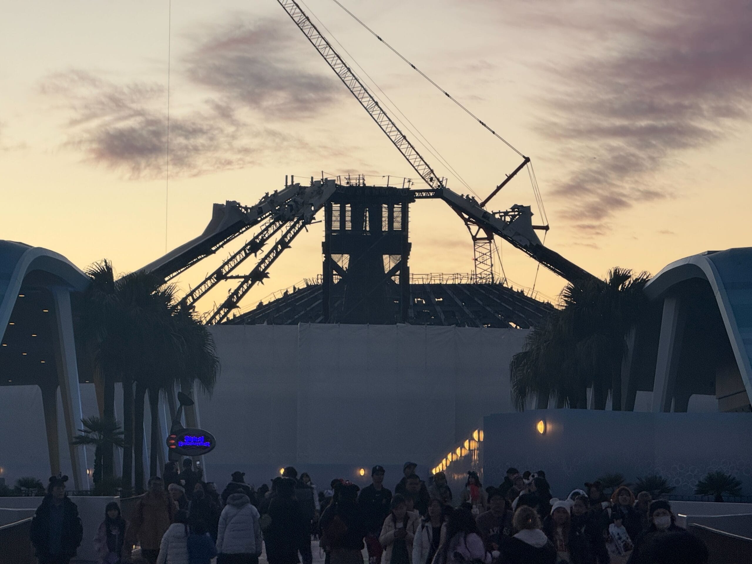 A large construction site with cranes is silhouetted against a sunset sky, surrounded by palm trees and a crowd of people walking.