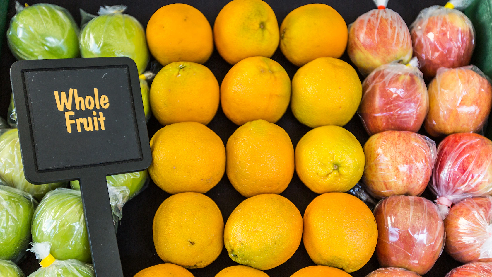 Oranges, green and red apples (in plastic) from Tropical Imports displayed with a sign labeled "Whole Fruit.