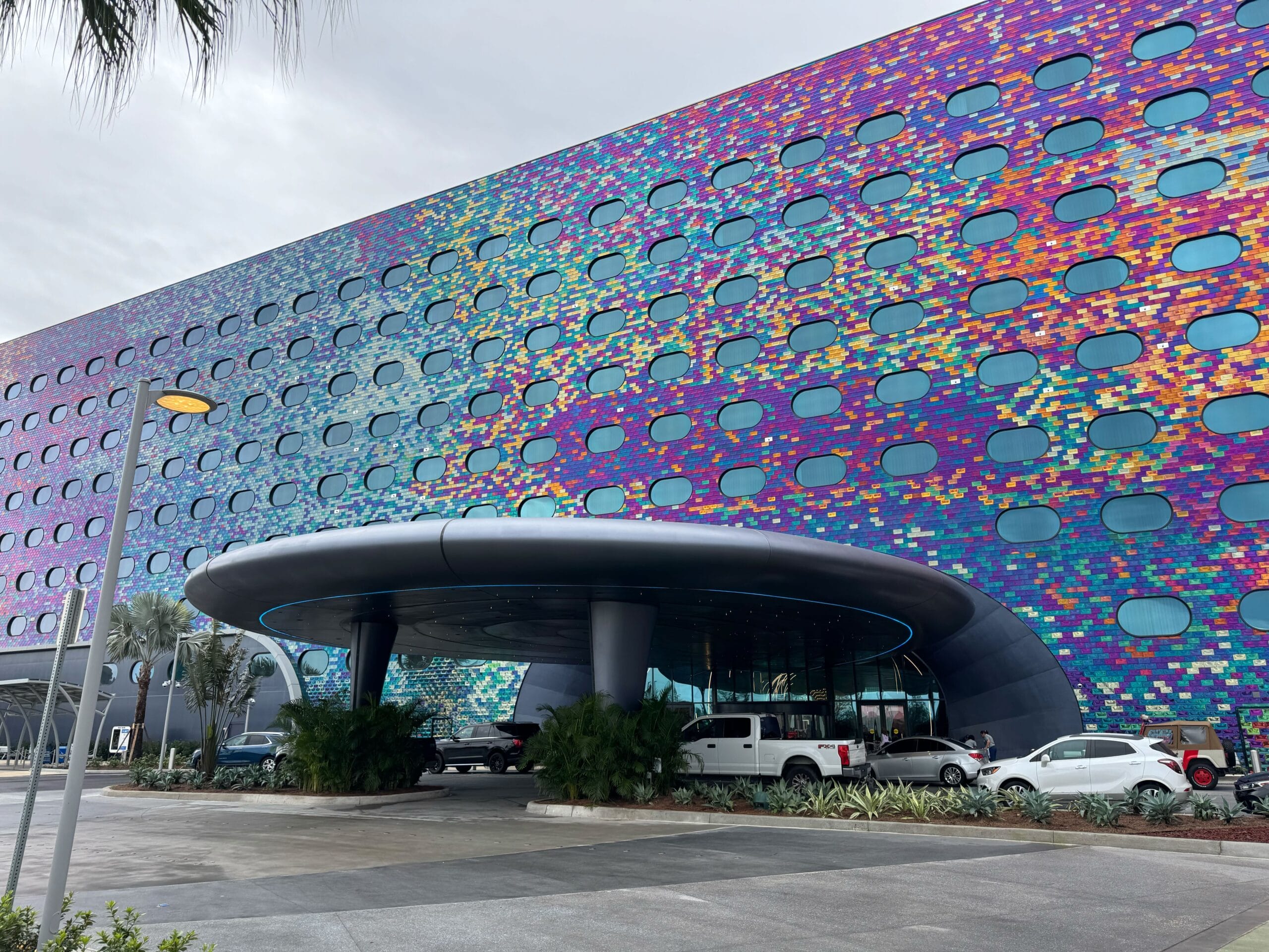 Colorful modern building facade with circular patterns above a round entrance. Several vehicles and palm trees are near the entrance. Sky is overcast.