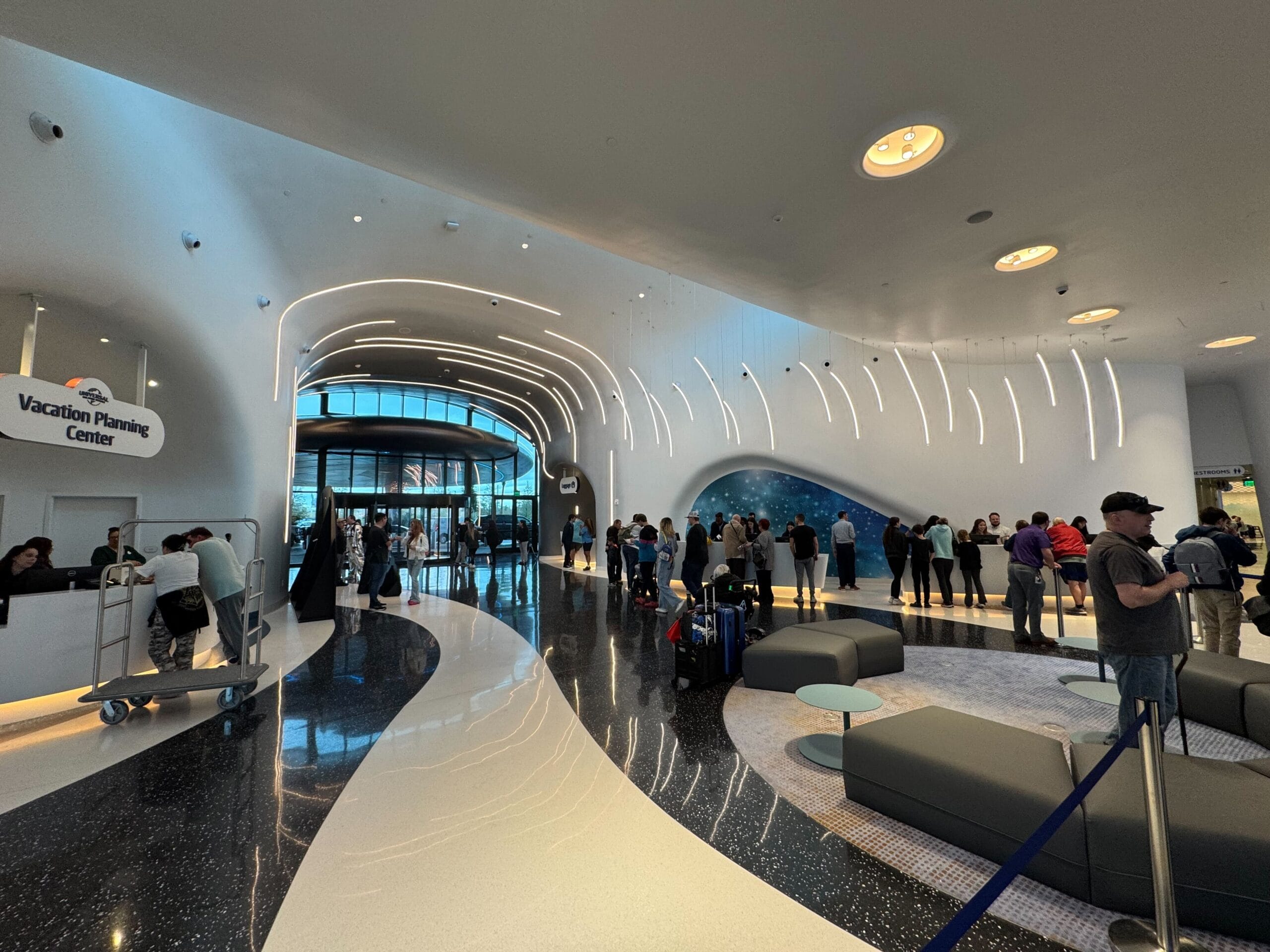 Modern lobby with curved architecture, shiny floors, and illuminated ceiling. People are checking in at counters and forming lines. Sign reads "Vacation Planning Center."