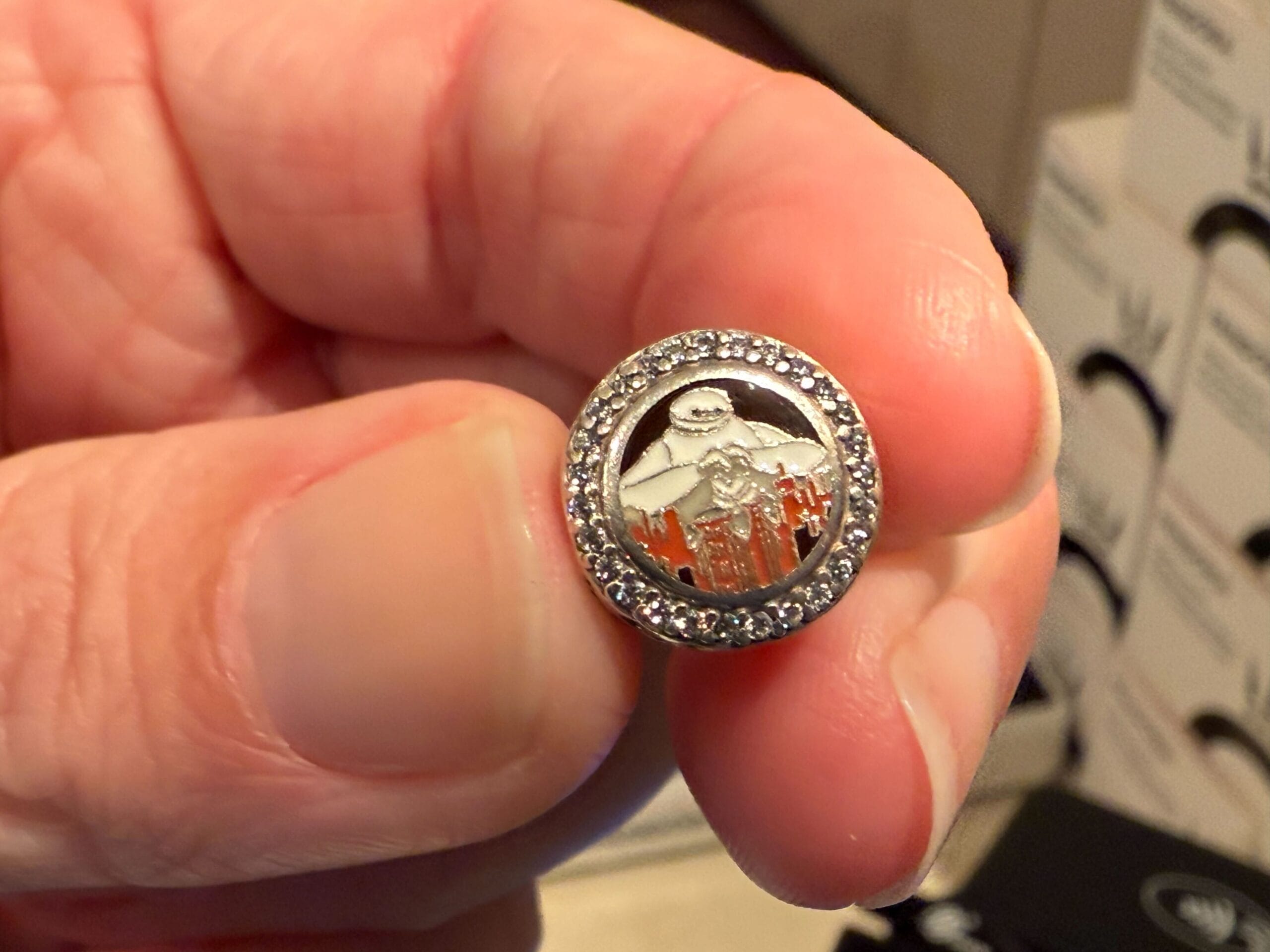 A hand holding a round, silver charm with a white ghost figure and sparkling rim.