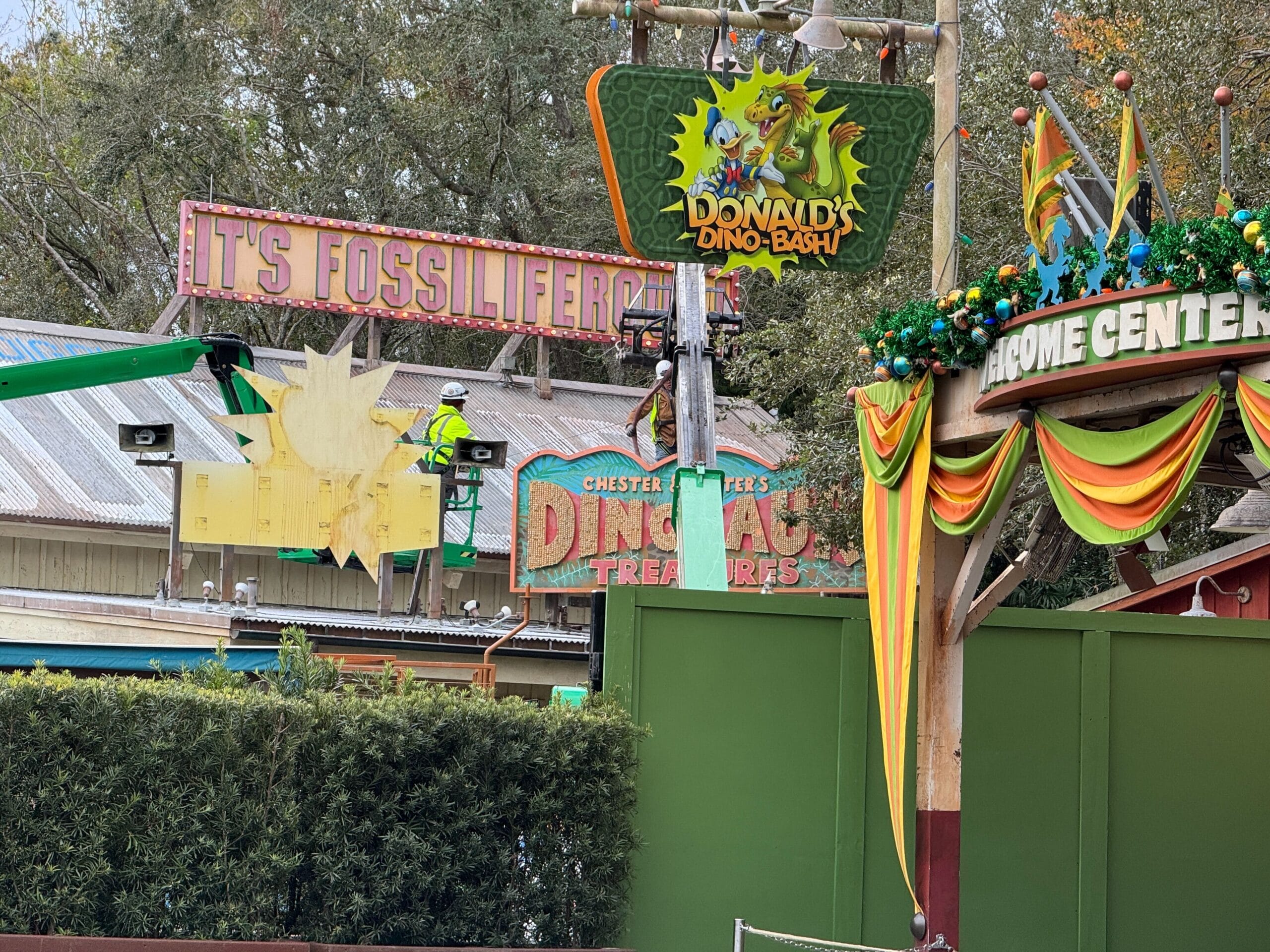 Workers on a lift install a DinoLand sign at the theme park attraction, surrounded by colorful decor and foliage.
