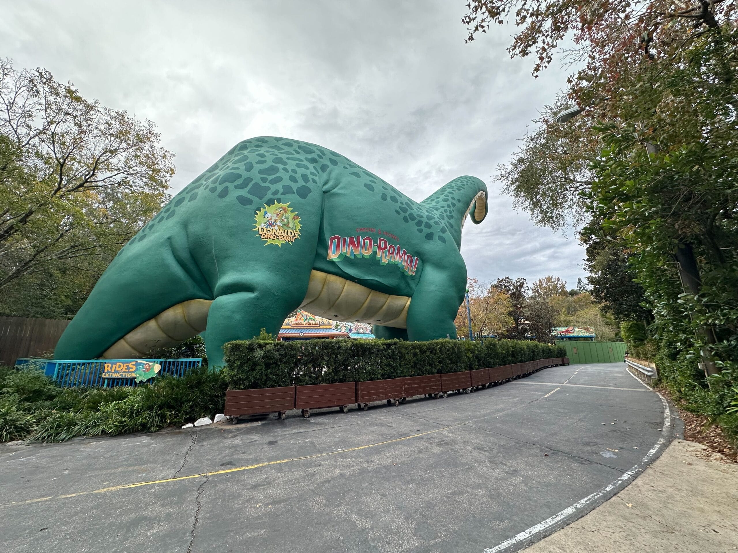 Nestled in the heart of Dinoland, a large green dinosaur structure towers majestically at an amusement park. With the "Dino Pond" sign gleaming nearby and surrounded by lush trees and bushes, a winding pathway invites visitors on an adventurous journey.