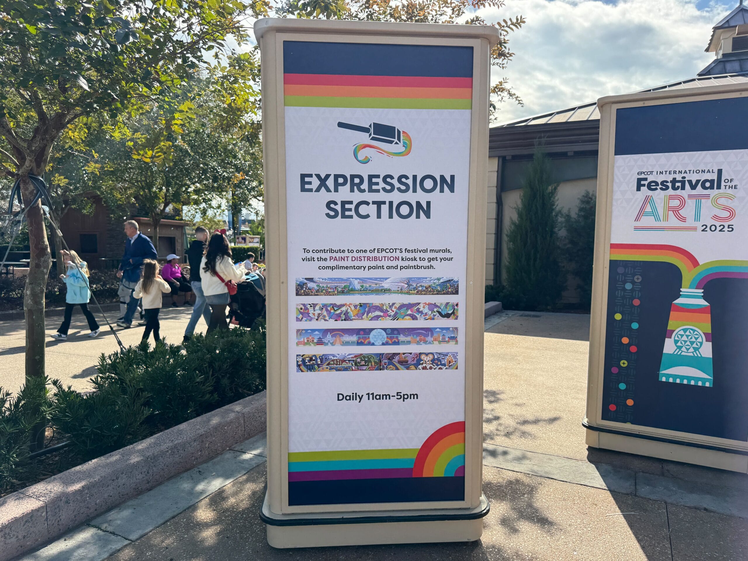 Sign for "Expression Section" at an arts festival. It invites participation in a paint-by-number activity available daily from 11am to 5pm. Nearby, people walk and trees provide shade.