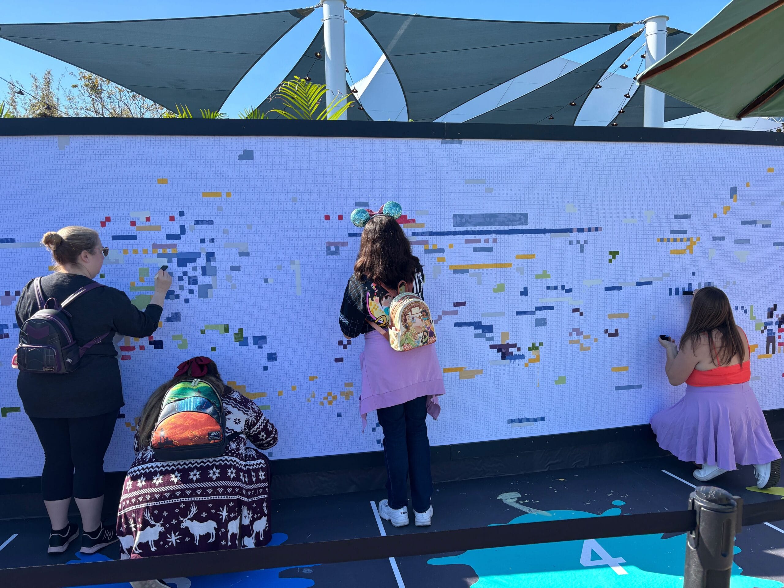 People placing mosaic tiles on a large outdoor art board, with colorful patterns starting to form in this vibrant expression of creativity. Each section comes alive as colors and shapes blend into a stunning visual symphony.