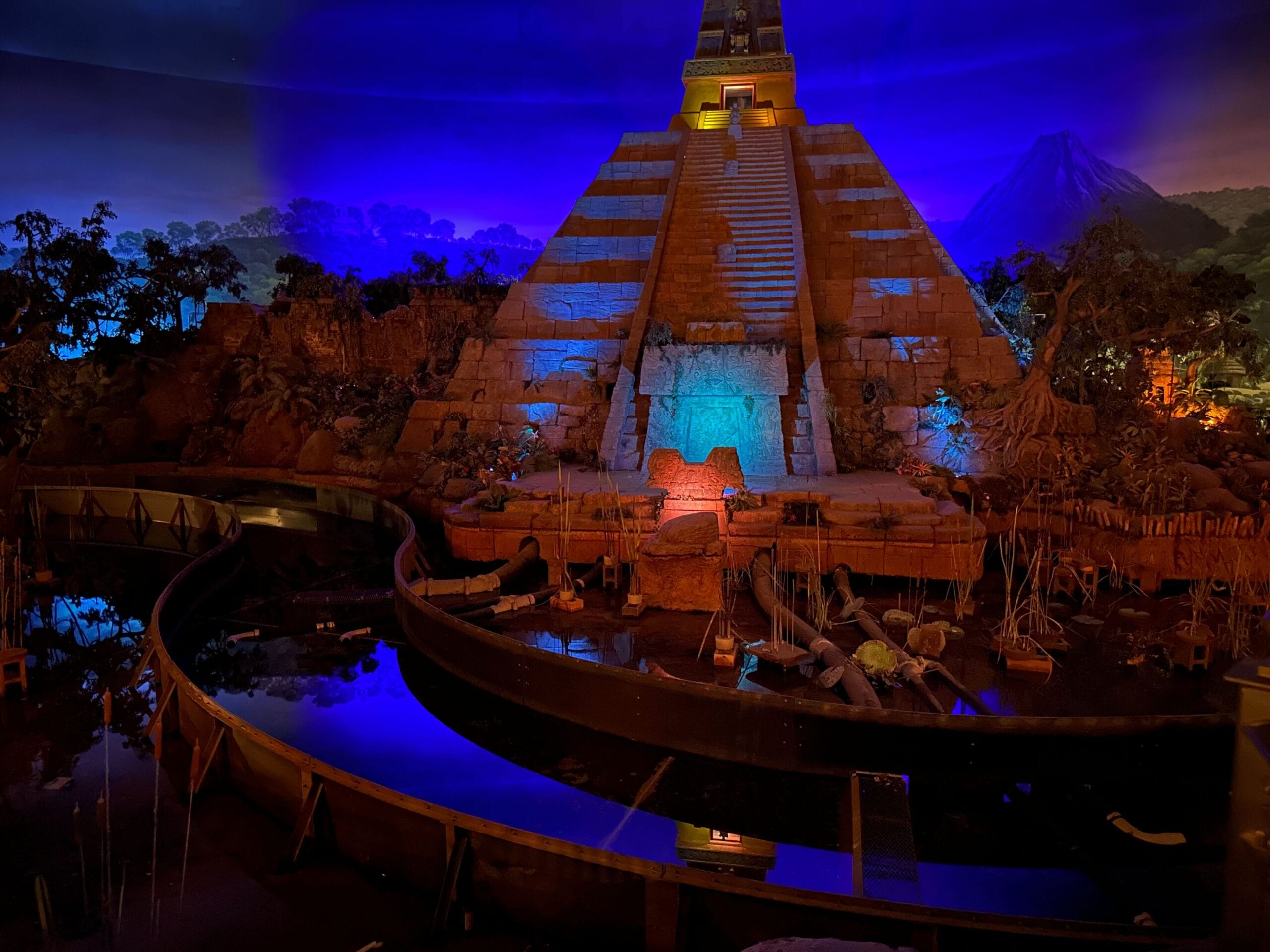A dimly lit pyramid with colorful lights projected on it, surrounded by water and vegetation, set against a dark blue sky.