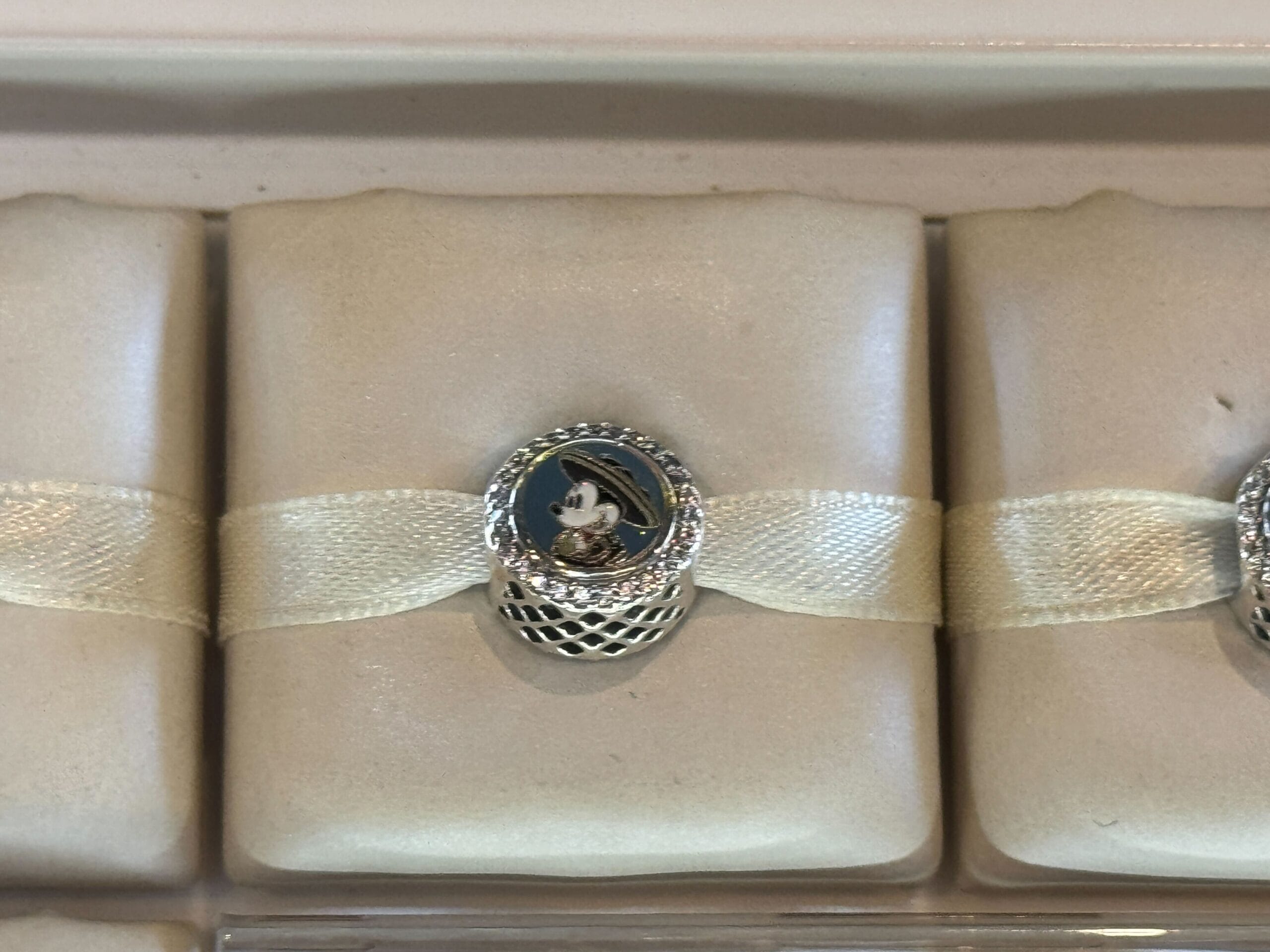 A silver charm depicting a cartoon mouse is secured on a beige ribbon, placed in a lined display case.