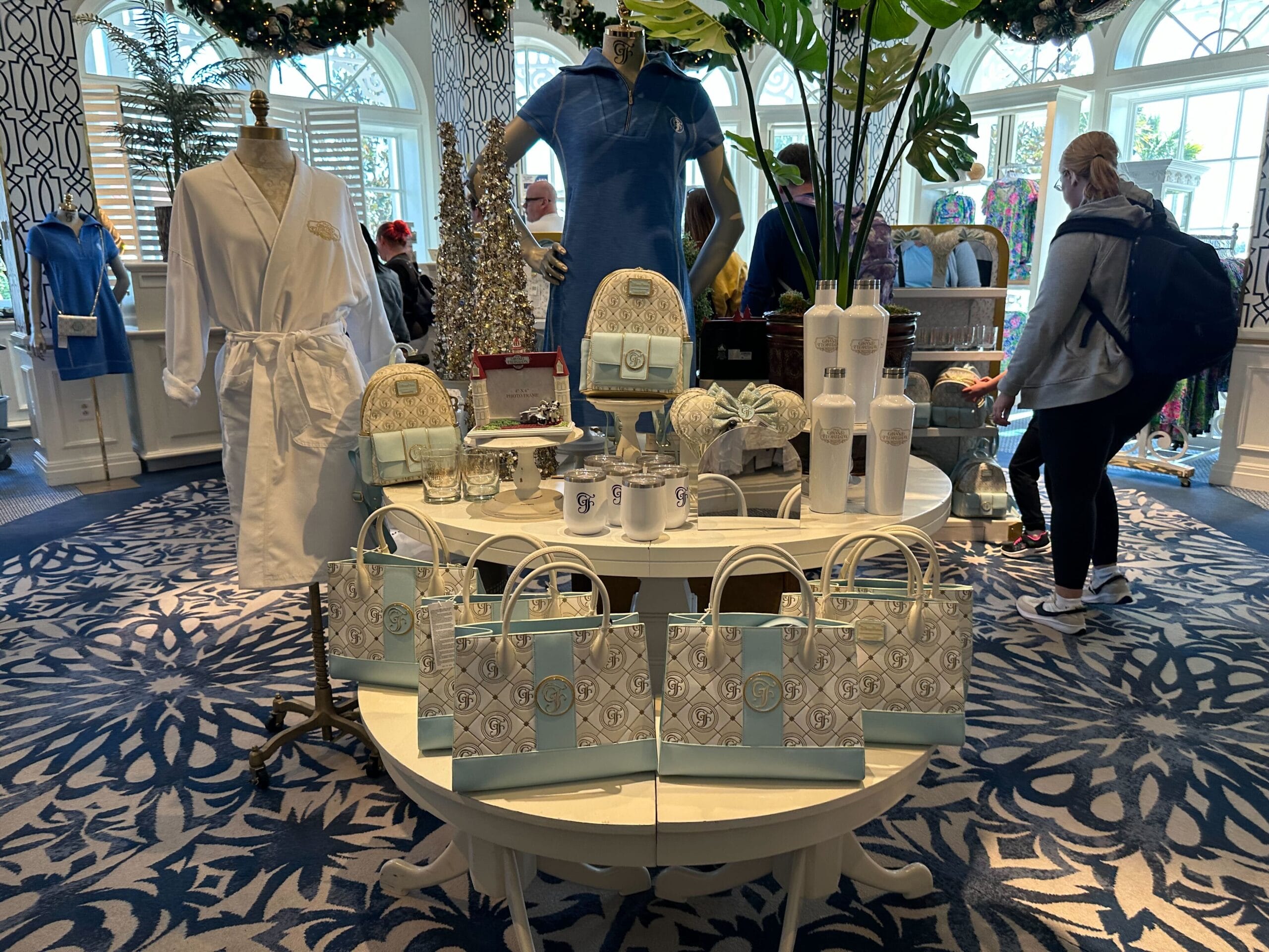 A retail display featuring bags, mugs, and backpacks on round tables, with mannequins wearing a white robe and a blue outfit.