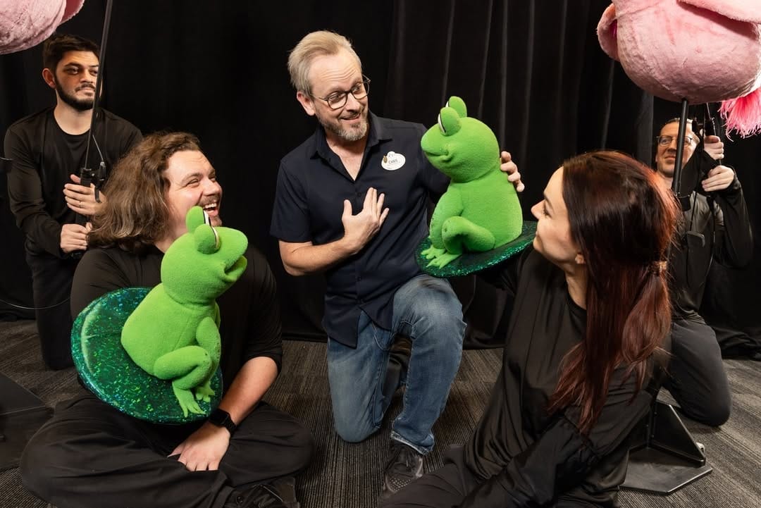 A group of people dressed in black are interacting with green frog puppets in a room with a black curtain.