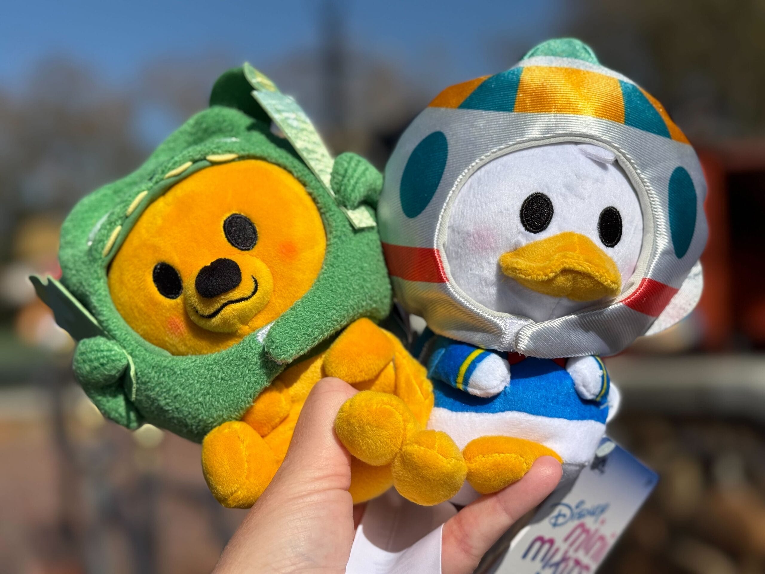 Two plush toys are held outdoors: one is a smiling yellow bear in a green outfit, the other is a white duck wearing a colorful outfit with blue and orange details.