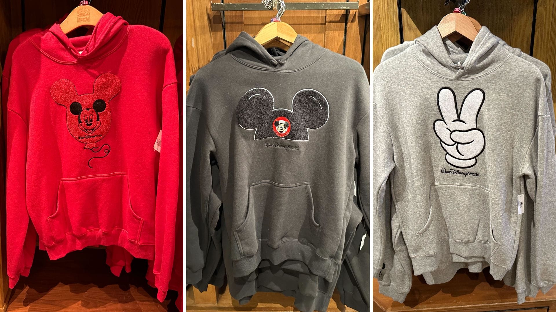 Three hoodies on hangers: a red one with a Mickey Mouse design, a gray featuring large Mickey ears, and a light gray adorned with a peace sign hand graphic. Perfect for fans, these Walt Disney World hoodies bring magic to casual wear.