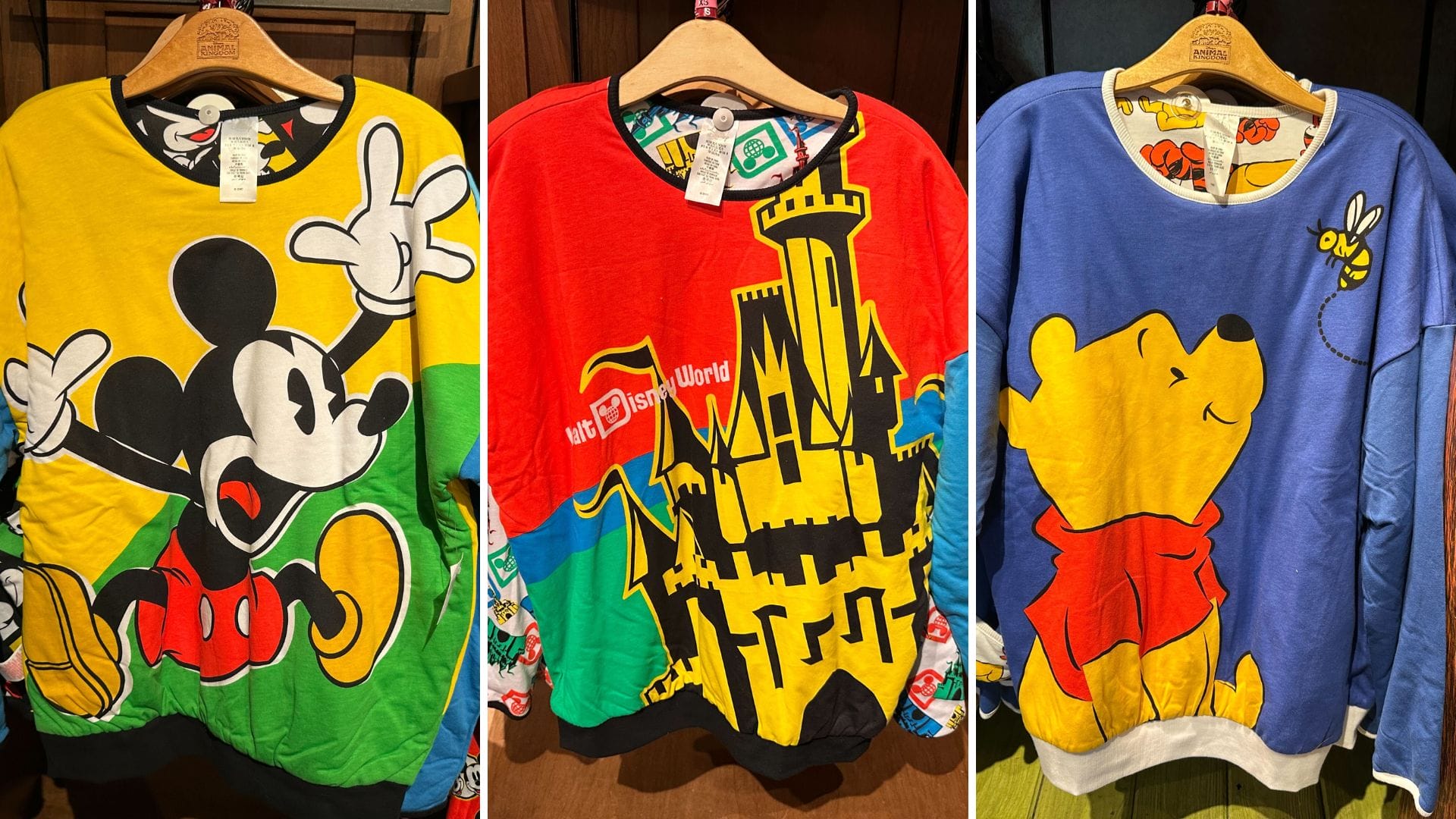 Three colorful sweatshirts on hangers featuring cartoon designs: left with Mickey Mouse, center with a castle and "Walt Disney World" text, right with Winnie the Pooh holding a balloon. The reversible pullover adds an extra touch of Disney magic to your wardrobe.