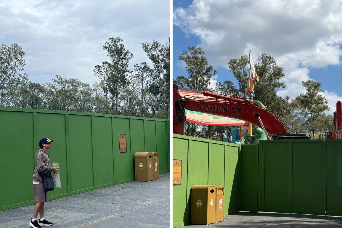 At a construction site with green barriers and equipment, a person stands outside the fence in casual clothing, holding items reminiscent of a Dino-Rama scene. Trees frame the backdrop against a cloudy sky.