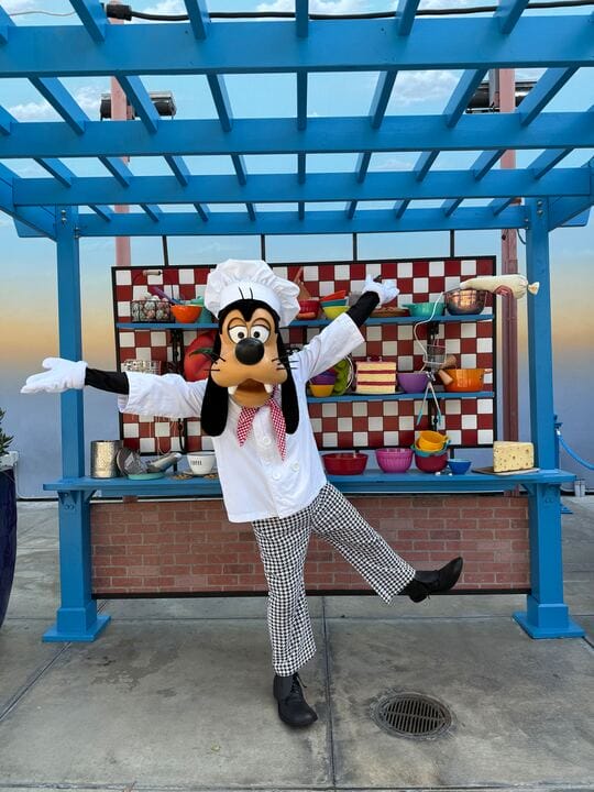 Under the blue pergola, Chef Goofy strikes a playful pose, surrounded by cooking props neatly arranged on shelves behind.