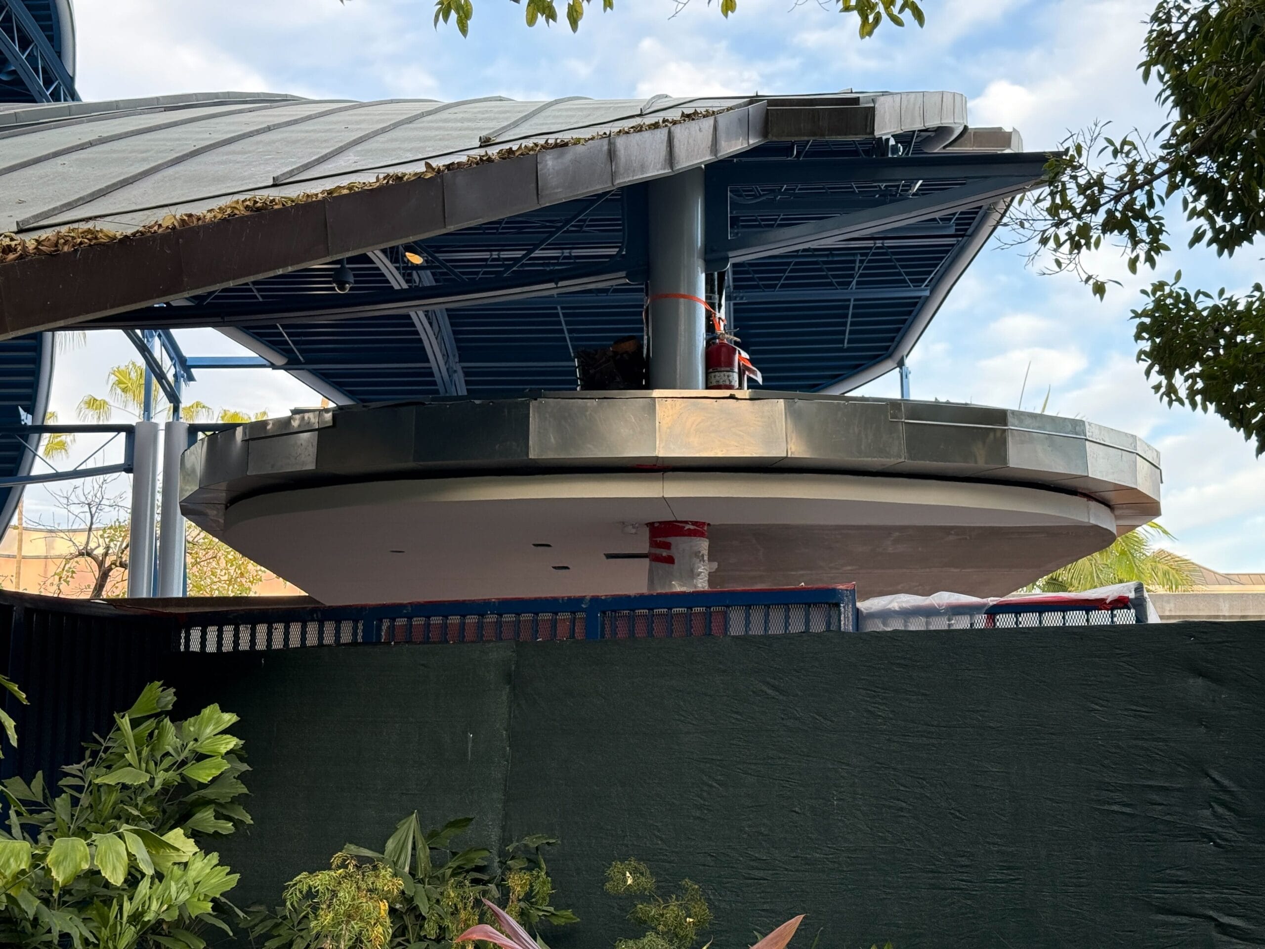 A construction site reveals a circular structure with a metal roof, resembling the beginnings of a Disneyland Monorail Station. Green fencing and lush foliage provide an enchanting foreground.