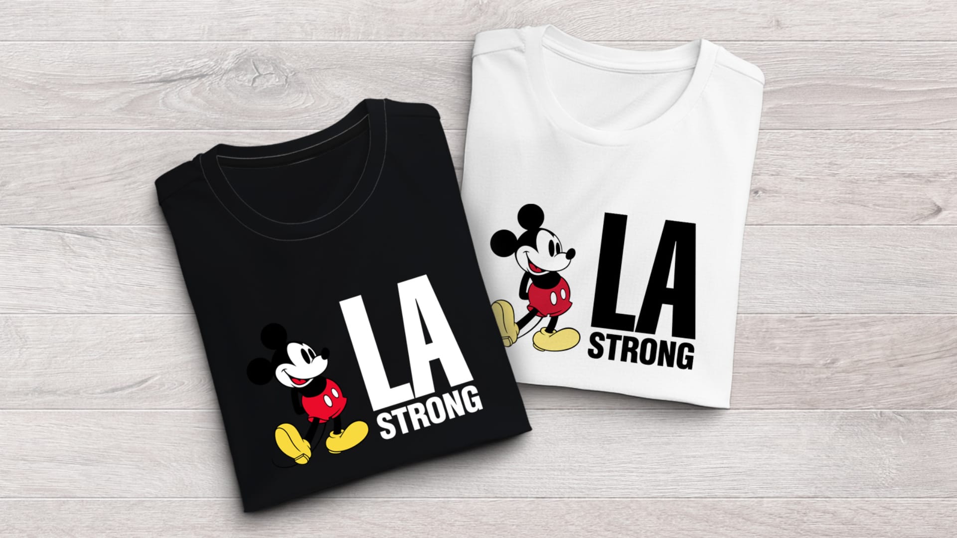 Two T-shirts on a wooden surface: one black, one white, both featuring cartoon mouse and text "LA STRONG" with city skyline silhouette.