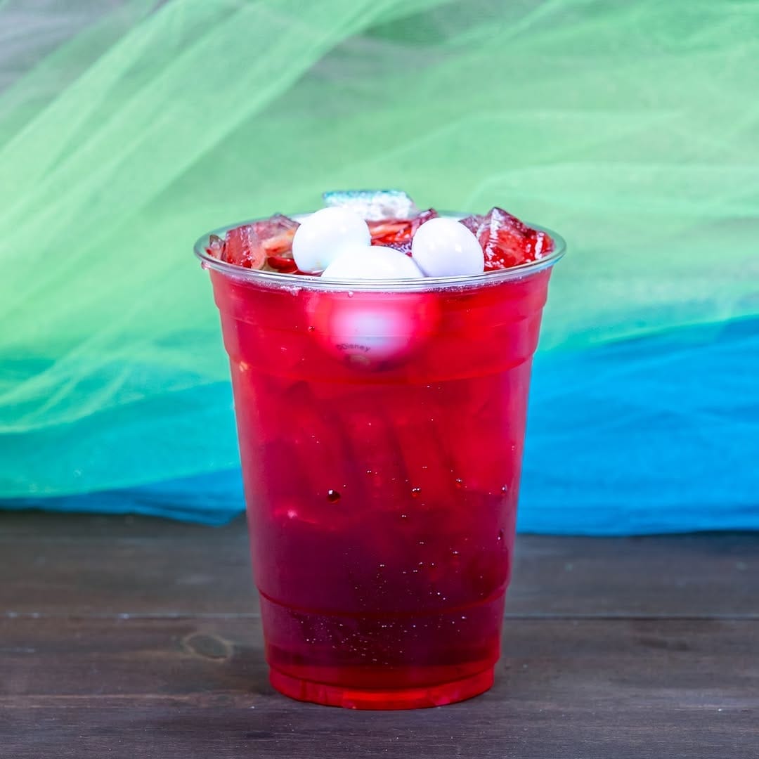 A red drink in a transparent plastic cup with ice and three round white garnish pieces, set against a colorful green and blue fabric background.