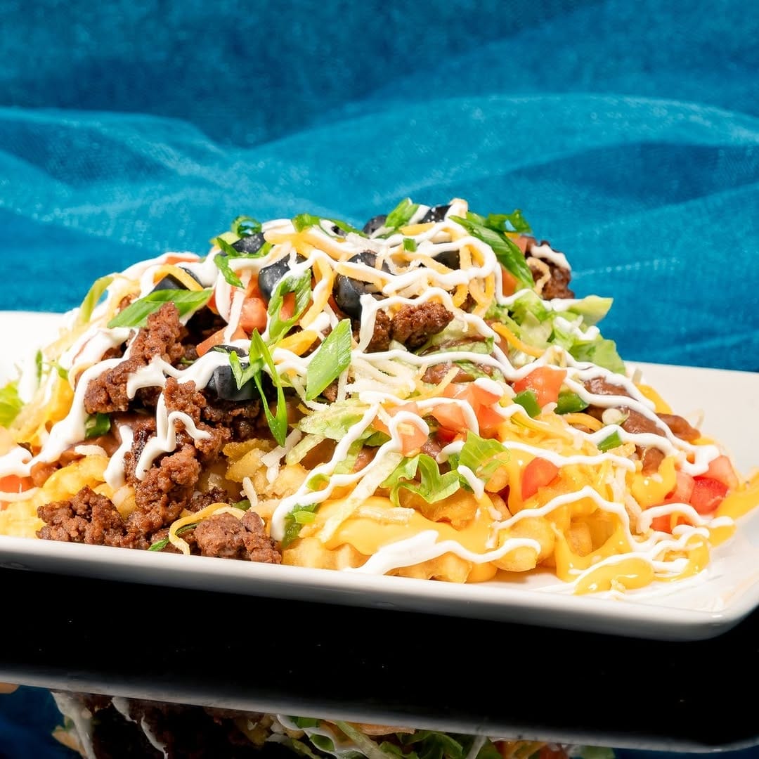 Loaded waffle fries topped with ground beef, shredded cheese, tomatoes, lettuce, olives, and drizzled with sauce, served on a white rectangular plate against a blue background.