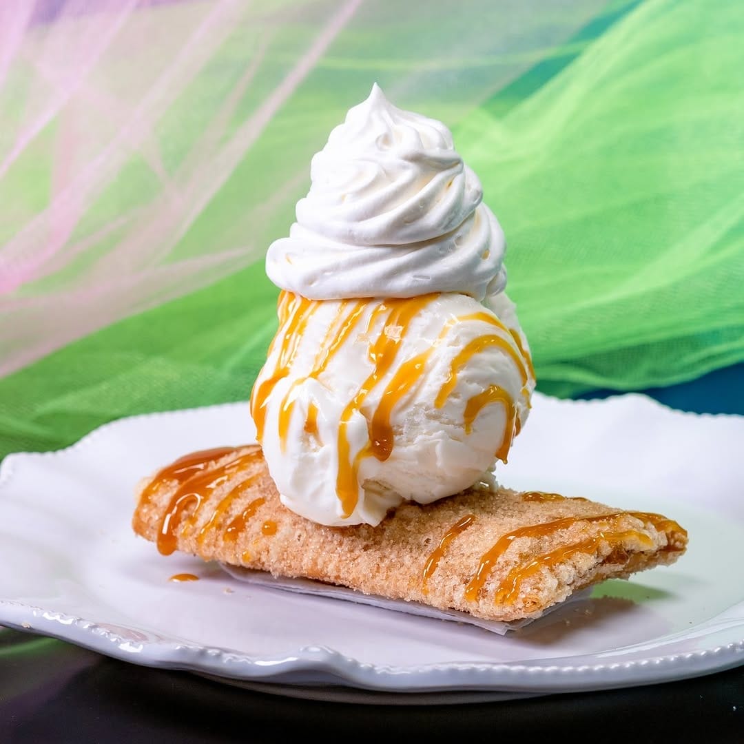 A dessert with ice cream topped with whipped cream and caramel sauce served on a pastry on a white plate. Green and pink fabric in the background.