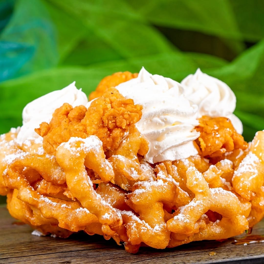 Funnel cake topped with fried chicken pieces, whipped cream, and powdered sugar on a wooden surface with a green blurred background.