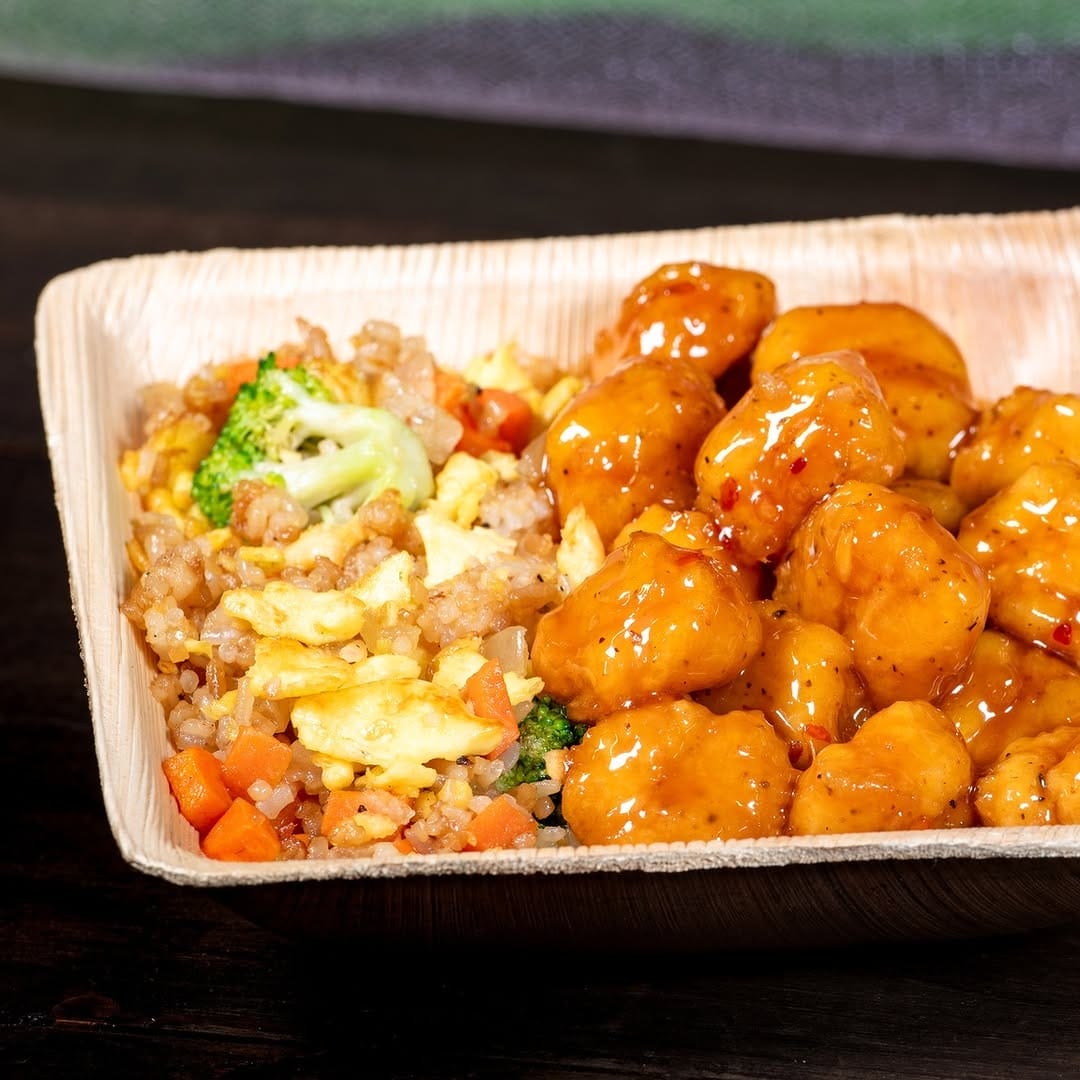 A wooden tray with orange chicken pieces and vegetable fried rice containing broccoli, carrots, and scrambled eggs.