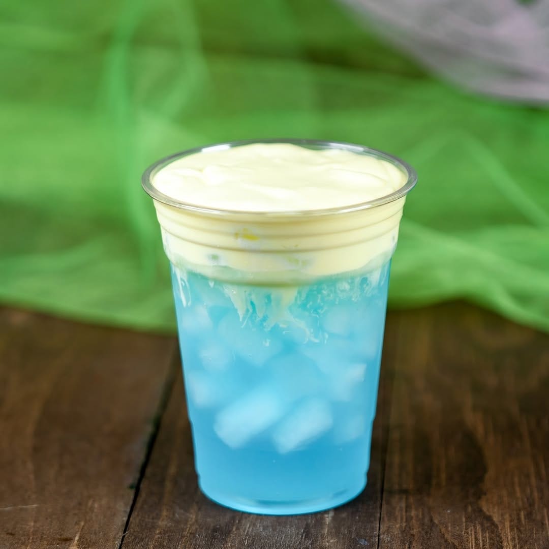 A glass of blue iced drink topped with a layer of creamy, yellow foam on a wooden surface, with a green background.