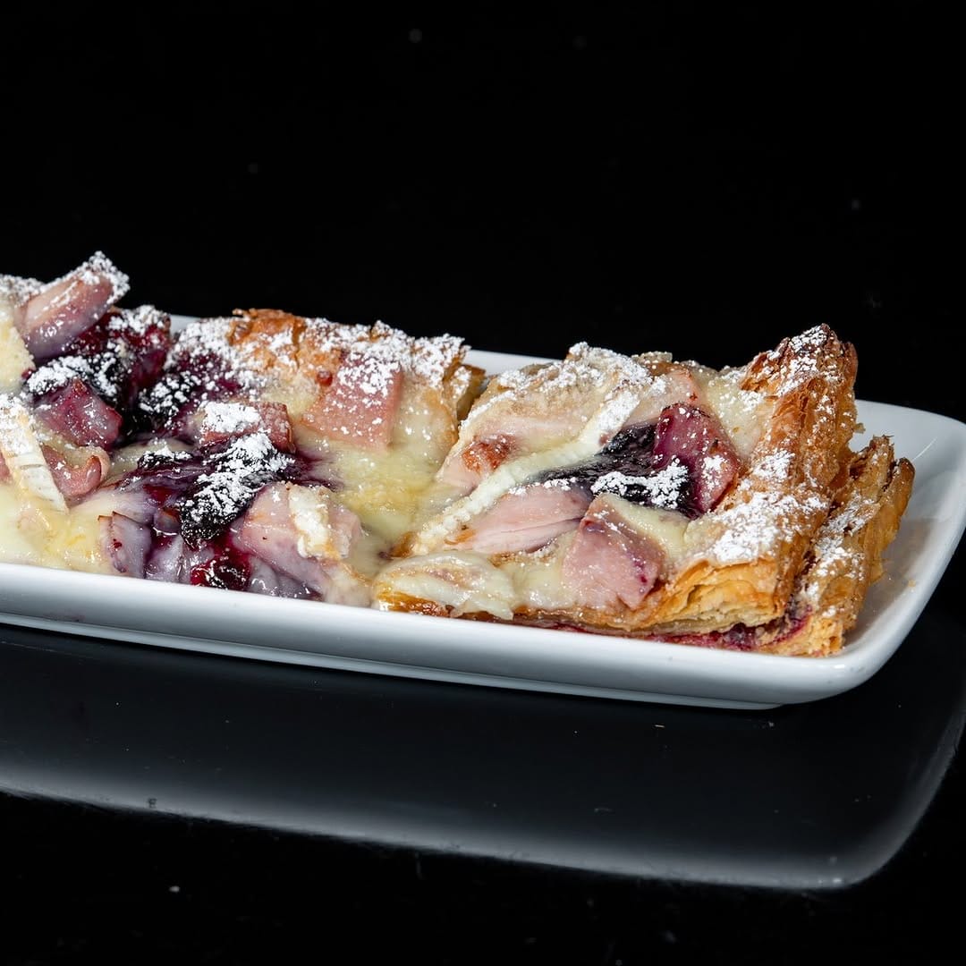 A rectangular pastry with fruit toppings and powdered sugar on a white plate against a black background.
