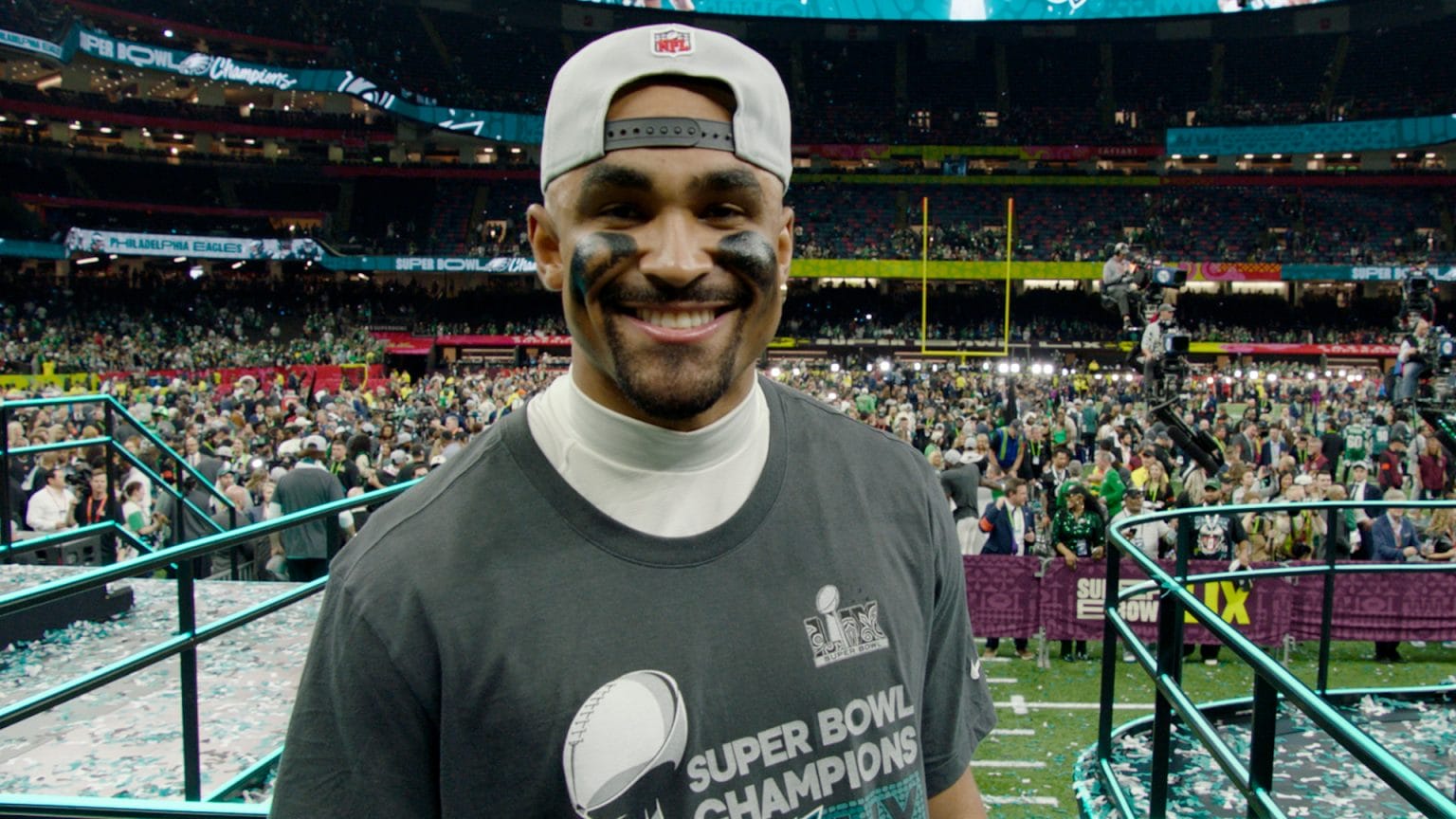 A football player proudly wearing a "Super Bowl Champions" shirt beams on the field during a vibrant celebration event, with colorful confetti scattered all around.