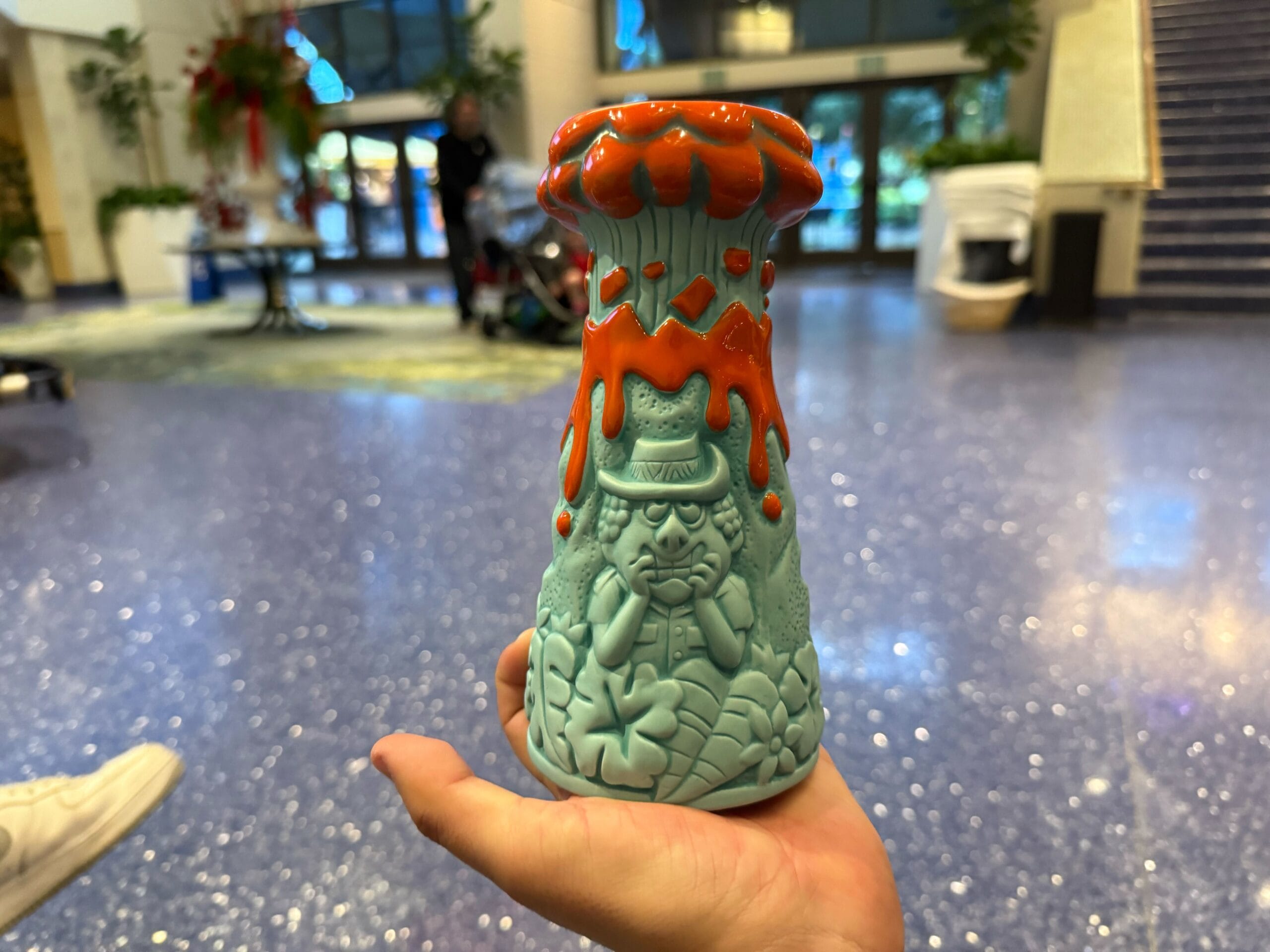 In a spacious, modern lobby with a shiny floor, a person holds an exquisite pottery vase with a sculpted face and red drips, reminiscent of the playful grooves found on a laughing lava mug.