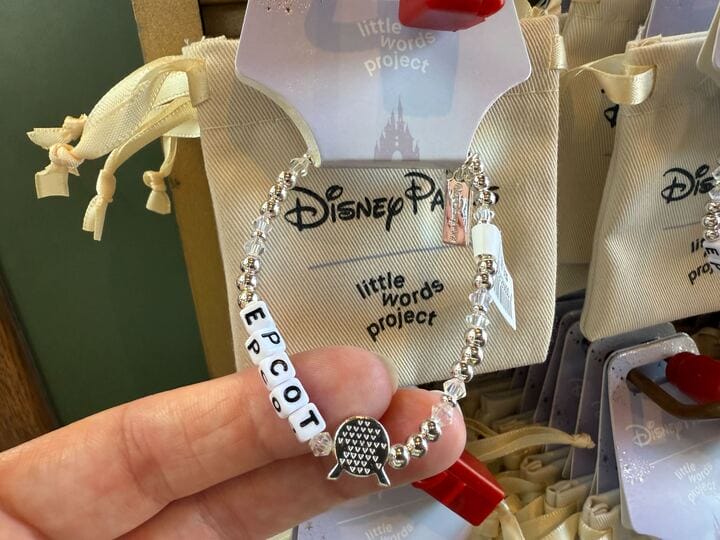 A hand holds a beaded bracelet with "Epcot" spelled out, featuring silver beads and a decorative charm, in front of a Disney Parks display.