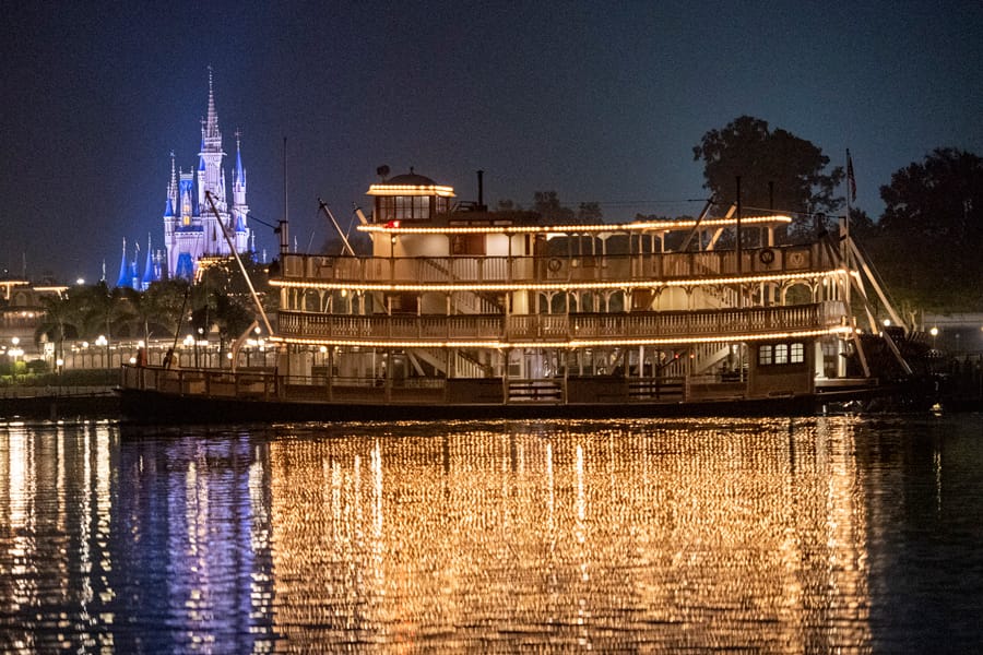 A Liberty Square Riverboat lit up at night with a glowing castle in the background, reflecting on the water.