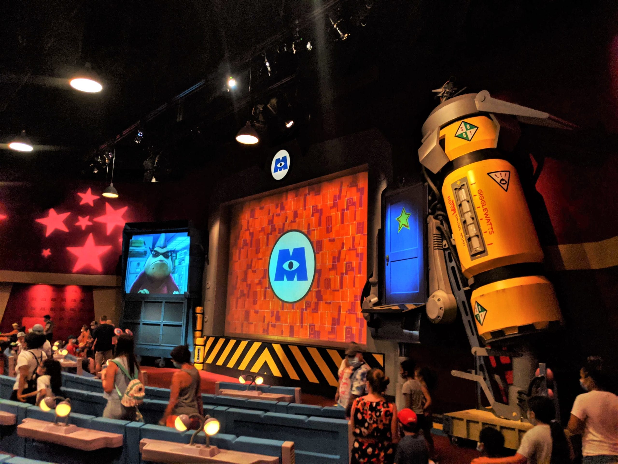 Theme park attraction room with Monsters Inc. Laugh Floor logo, large yellow rocket, and people seated inside.